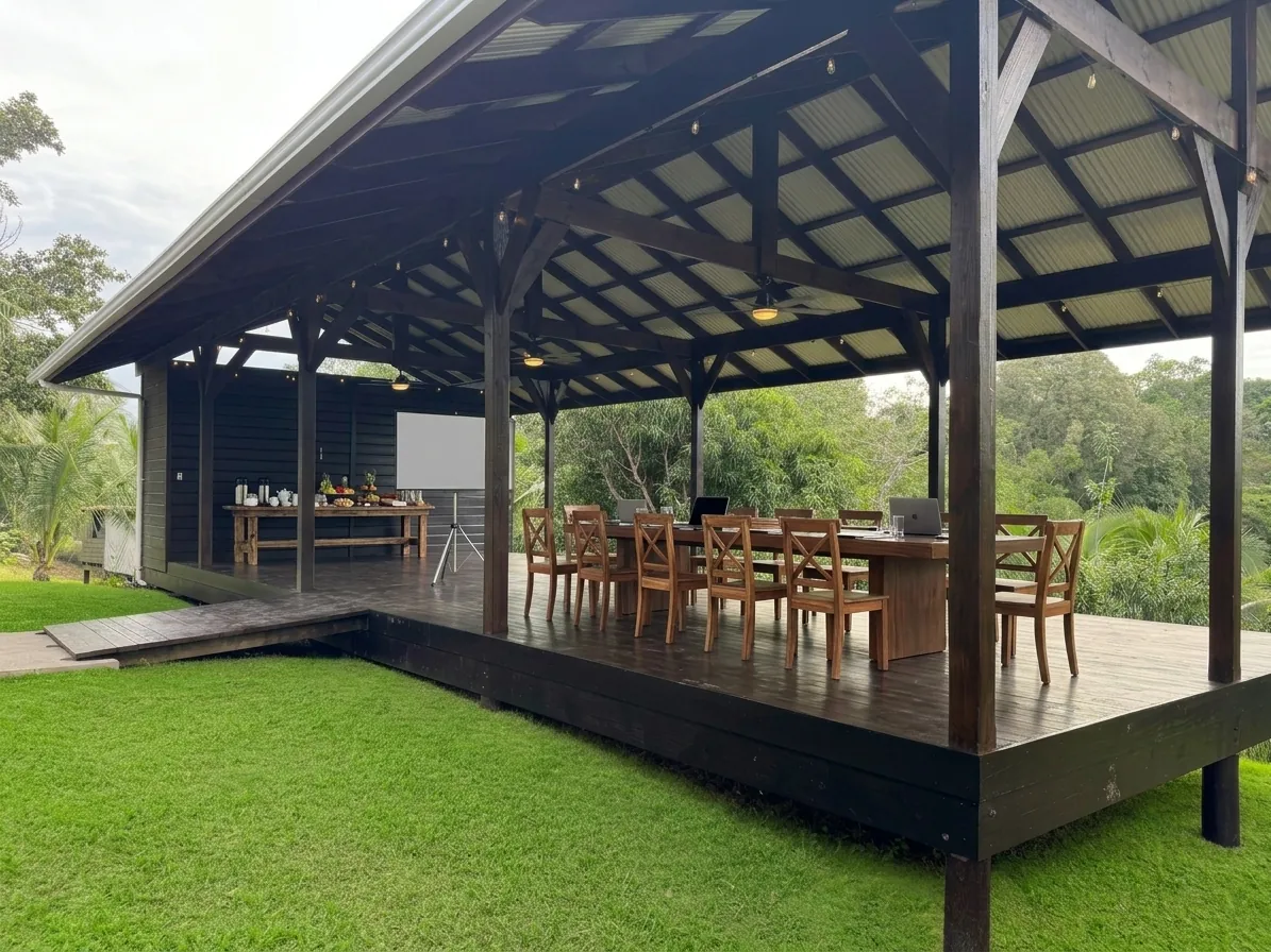 The pavilion in daylight set up for a meeting, with a projector screen on a tripod, laptops on the long table and breakfast on a side table